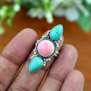Pode incluir: Um anel de prata com uma pedra rosa redonda central ladeada por duas pedras turquesas em forma de gota. A cravação inclui detalhes decorativos de contas de prata e fio torcido que rodeiam as pedras.
