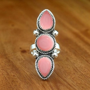 Puede incluir: Un anillo de plata con tres piedras lisas de color rosa claro dispuestas verticalmente. La piedra central es circular, mientras que las de arriba y abajo tienen forma de lágrima. Cada piedra está enmarcada por un borde de cuerda de plata trenzada y pequeñas cuentas.
