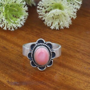 May include: A silver ring featuring a smooth, oval-shaped pink stone set in the centre. The stone is surrounded by a twisted rope-like metal border and a scalloped, petal-shaped frame. The band of the ring is plain, polished silver metal.
