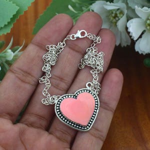 May include: A silver chain necklace featuring a heart-shaped pendant with a smooth, light pink center inlay. The pendant is framed by a textured, beaded silver border, and the necklace is secured with a lobster claw clasp closure.