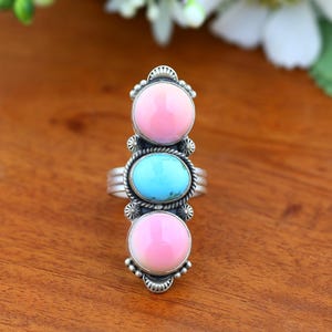 May include: A silver ring featuring three vertically aligned cabochon stones. The top and bottom stones are round and light pink, while the center stone is oval and bright turquoise. The silver setting includes decorative beaded metal accents.