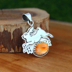 May include: A silver pendant featuring a leaping rabbit silhouette positioned above a round, orange, textured stone set in a serrated metal bezel. The pendant includes a silver bail at the top for attachment to a chain or cord necklace.