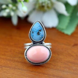 May include: A silver ring featuring two stacked stones: a pear-shaped blue turquoise stone with dark matrix patterns at the top and a smooth, oval-shaped light pink stone at the bottom. Both stones are set in twisted silver rope borders.