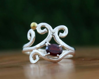 Puede incluir: Un anillo de plata con un diseño en espiral, con una piedra preciosa ovalada de color rojo intenso y un pequeño acento dorado. El anillo se encuentra sobre un fondo verde y marrón borroso.