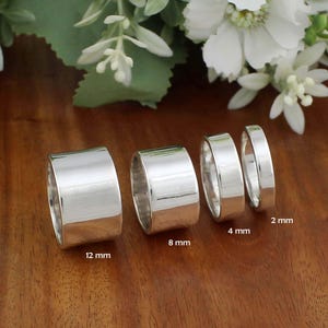 Puede incluir: Se muestran cuatro anillos de plata pulida de diferentes anchos. Los anillos miden 12 mm, 8 mm, 4 mm y 2 mm de ancho. Los anillos reflejan la luz y están colocados sobre una superficie de madera. Flores blancas y hojas verdes en el fondo.