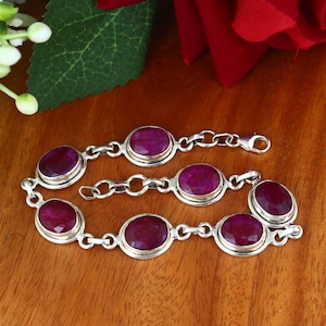May include: A silver bracelet featuring oval-cut, deep red gemstones. The bracelet has a delicate chain design with seven gemstones. The clasp is visible, and the bracelet is displayed on a wooden surface.