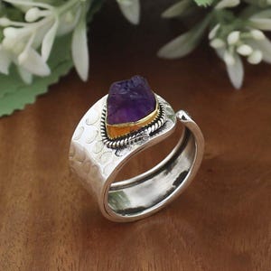 Op de afbeelding: Een zilveren ring met een ruwe, paarse amethist edelsteen gezet in een goudkleurige bezel. De ring heeft een getextureerde band met een gehamerd ontwerp. De edelsteen is dieppaars.