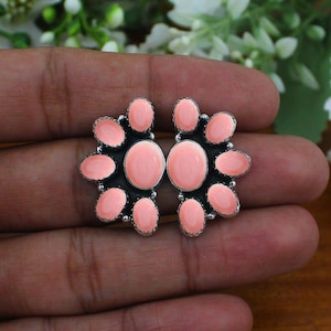 May include: A pair of stud earrings featuring oval-shaped, light pink stones set in dark, oxidized silver. Each earring is arranged in a cluster pattern with a larger central stone surrounded by smaller oval stones in a fan-like formation.
