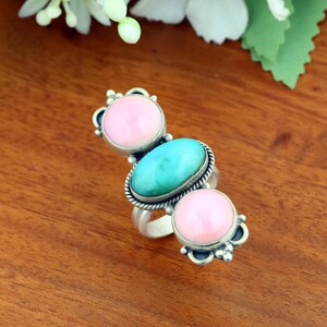 Peut inclure: Une bague en argent ornée d'une pierre turquoise ovale centrale flanquée de deux pierres roses rondes. La monture présente des détails en métal argenté avec une bordure en corde torsadée autour de la pierre centrale et des perles d'argent.