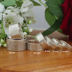 May include: A collection of silver rings with a repeating wave pattern. The rings vary in width from 2 mm to 14 mm. The rings are displayed on a wooden surface with white flowers and a red rose in the background.