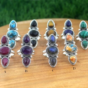 May include: A collection of silver rings with various gemstone settings. The rings feature oval and teardrop-shaped stones in shades of red, purple, blue, green, and brown. The gemstones are set in ornate silver bands.