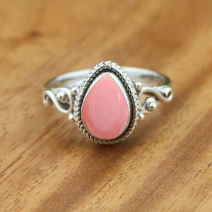 Op de afbeelding: Een zilveren ring met een centrale druppelvormige roze steen. De steen is omringd door een gedraaide, touwachtige metalen rand. De ringband vertoont decoratieve kruldetails aan beide zijden van de centrale zetting.