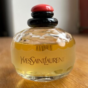 Back of vintage Yves Saint Laurent Paris perfume bottle placed on a warm wooden surface, highlighting the label’s text and the pale yellow perfume inside. The black and red cap contrasts with the wood grain