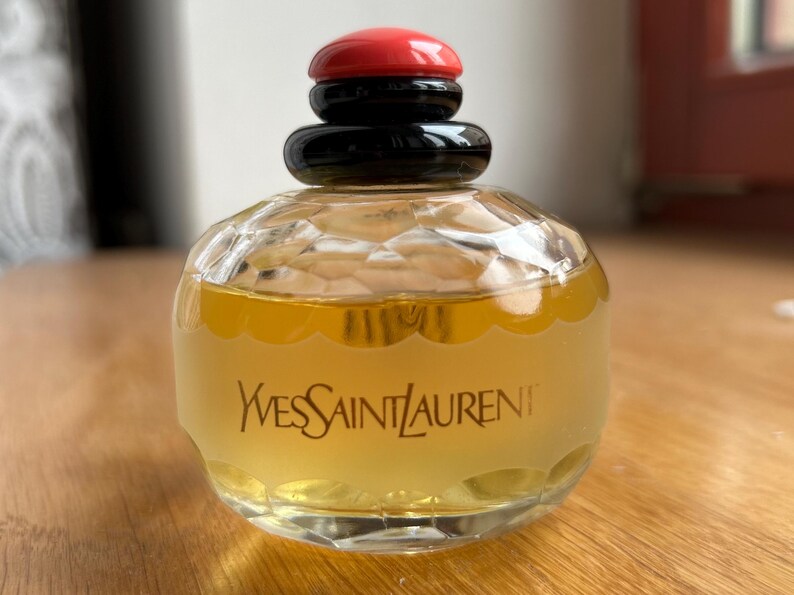 Back of vintage Yves Saint Laurent Paris perfume bottle placed on a warm wooden surface, highlighting the label’s text and the pale yellow perfume inside. The black and red cap contrasts with the wood grain