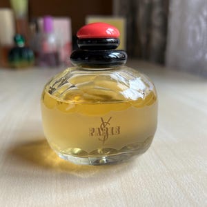Front of vintage Yves Saint Laurent Paris perfume bottle on a neutral surface. The rectangular clear glass bottle holds pale yellow liquid and has a black and red cap. The elegant label is visible on the front