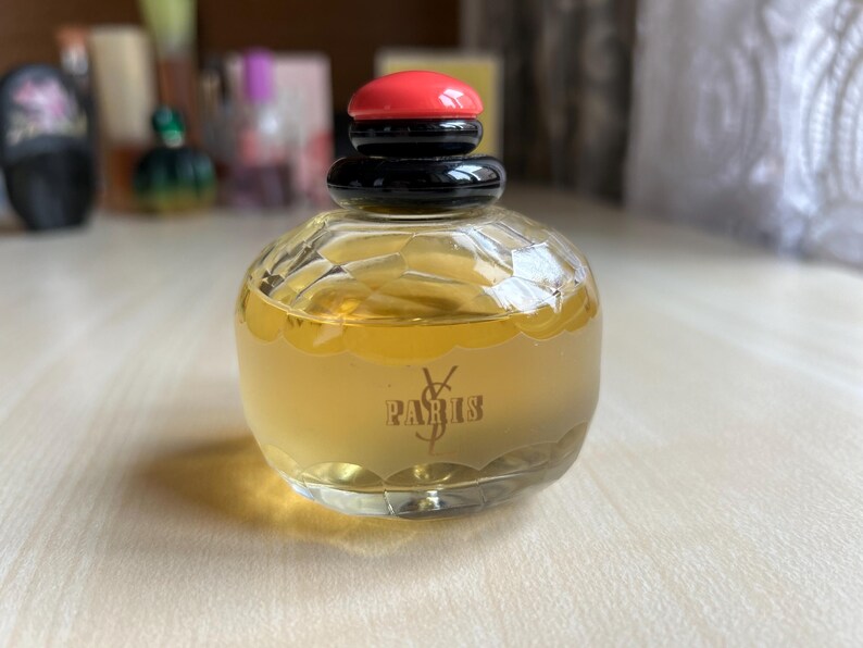 Front of vintage Yves Saint Laurent Paris perfume bottle on a neutral surface. The rectangular clear glass bottle holds pale yellow liquid and has a black and red cap. The elegant label is visible on the front