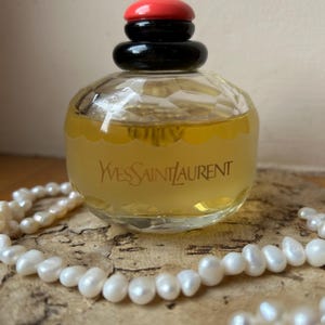 Back of vintage Yves Saint Laurent Paris perfume bottle resting on cork with scattered pearls around it, showcasing the label details and pale yellow liquid inside. The black and red cap tops the bottle.