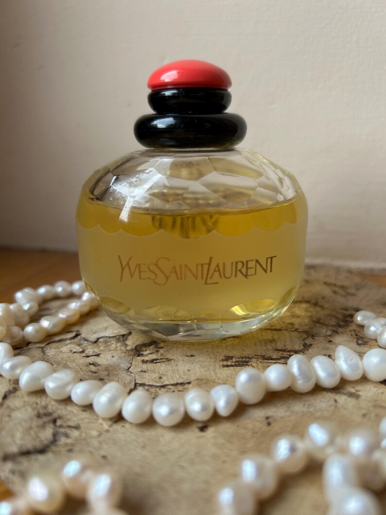 Back of vintage Yves Saint Laurent Paris perfume bottle resting on cork with scattered pearls around it, showcasing the label details and pale yellow liquid inside. The black and red cap tops the bottle.