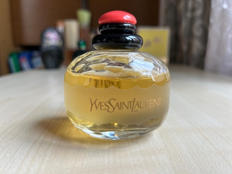 Back of vintage Yves Saint Laurent Paris perfume bottle on a neutral surface, showing the text Yves Saint Laurent details on the bottle. The pale yellow liquid and black and red cap are visible