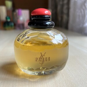 Front of vintage Yves Saint Laurent Paris perfume bottle on a neutral surface. The rectangular clear glass bottle holds pale yellow liquid and has a black and red cap. The elegant text is visible on the front