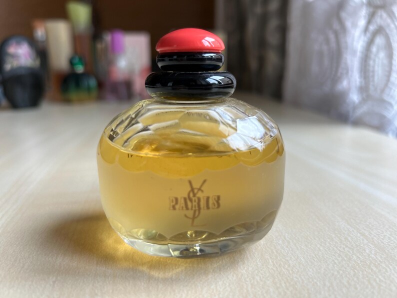 Front of vintage Yves Saint Laurent Paris perfume bottle on a neutral surface. The rectangular clear glass bottle holds pale yellow liquid and has a black and red cap. The elegant text is visible on the front