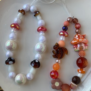 Puede incluir: Dos colgantes de cuentas con diseños de setas y bosque. Un colgante presenta cuentas blancas, iridiscentes y marrones con acentos de setas. El otro colgante tiene una ardilla, una flor y una casa de setas con cuentas naranjas, marrones y rosas.