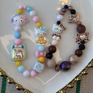 May include: Two beaded bracelets featuring Hello Kitty charms and colorful beads. One bracelet has a backpack charm, while the other has a heart-shaped charm. The beads are in shades of pink, blue, yellow, and brown.