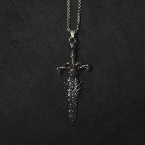 以下が含まれることがあります： 詳細な剣のデザインが施されたシルバーカラーのペンダントネックレス。剣のペンダントには複雑な彫刻が施され、お揃いのチェーンから吊り下げられています。背景は無地の黒です。