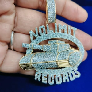 Könnte beinhalten: Goldfarbener Anhänger mit den Worten "NO LIMIT RECORDS" und einem Panzer-Design, besetzt mit kleinen, funkelnden Steinen. Der Anhänger hat eine passende Öse, das Design vor blauem Hintergrund.