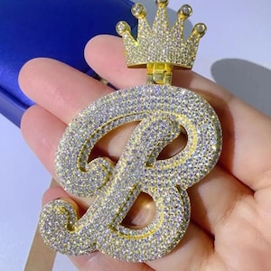 Może przedstawiać: Złoty wisiorek w kształcie litery "B", inkrustowany licznymi małymi, błyszczącymi kamieniami. Mała korona znajduje się na szczycie litery. Wisiorek jest trzymany w dłoni, prezentując jego skomplikowany design i luksusowy wygląd.