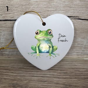 May include: A white heart-shaped ornament with a watercolor illustration of a green frog. The frog has large eyes and is centered on the ornament. The text "Dein Frosch" is written in black script. A gold string is attached for hanging.