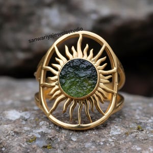 Könnte beinhalten: Ein goldfarbener Ring mit einem Sonnenmuster. Der Ring hat einen kreisförmigen, dunkelgrünen Stein in der Mitte. Die Sonnenstrahlen erstrecken sich nach außen und erzeugen einen auffälligen visuellen Effekt. Der Ring ist vor einem strukturierten, grauen Hintergrund platziert.