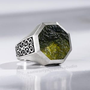 May include: A silver ring featuring a large, irregularly shaped green gemstone. The ring has a geometric design around the stone and decorative scrollwork on the band. The ring is set against a bright, reflective surface.