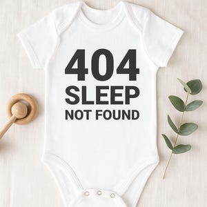 Könnte beinhalten: Weißer Baby-Body mit dem schwarzen Text "404 SLEEP NOT FOUND". Der Body hat kurze Ärmel und Druckknöpfe am unteren Ende. Ein Holzspielzeug und ein Zweig mit Grünzeug sind im Hintergrund.