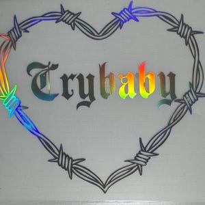 May include: A heart-shaped design made of barbed wire. The word "Trybaby" is written inside the heart in a gothic font. The text and some parts of the barbed wire have a rainbow holographic effect.