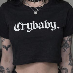 May include: Black crop top with the word "Crybaby" in white gothic font. The model is wearing a black studded choker and a silver padlock necklace. Tattoos are visible on the arms.