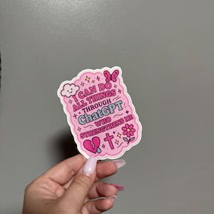 May include: A pink sticker with the text "I CAN DO ALL THINGS THROUGH ChatGPT WHO STRENGTHENS ME." The sticker features a cloud, butterfly, broken heart, cross, and flower illustrations. The sticker is held by a hand with long, pink nails.