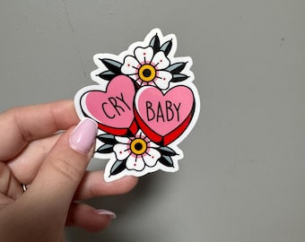 May include: A sticker featuring two pink heart-shaped candies with the words "CRY" and "BABY" in black. The hearts are surrounded by white flowers with yellow centers and black leaves. The sticker is held against a gray background.