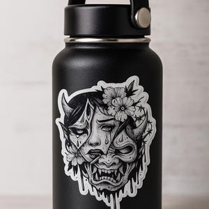 May include: Black insulated water bottle with a black handle and a silver ring. A black and white sticker of a crying woman and a demon face with flowers and horns is on the bottle.