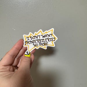 Può includere: Un adesivo bianco con testo nero che recita "I DON'T WANT SPONSERBILERTIES NO MORE" in una forma a stella gialla e nera. L'adesivo è tenuto in mano con un'unghia gialla con un disegno a palla 8 nera.
