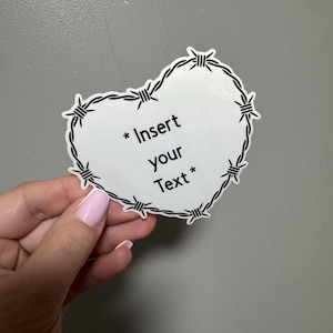 May include: A white heart-shaped sticker with a black barbed wire border. The sticker has the text "* Insert your Text *" in black. The sticker is held by a hand with a pink painted nail.