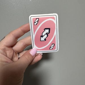 May include: A pink and white Uno reverse card. The card features a pink background with a black and white reverse arrow symbol in the center, and smaller reverse arrow symbols in each corner. The card has a white border.