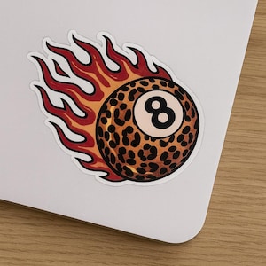 May include: A sticker featuring a leopard-print eight-ball engulfed in flames. The flames are red and orange with black outlines. The eight-ball has a white number eight.