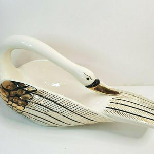 May include: A white ceramic swan-shaped dish with gold and black accents. The swan has a long, curved neck and a detailed feather design on its wings. The beak is gold, and the eye is black.