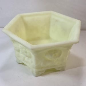 May include: A pale yellow, hexagonal-shaped planter with a textured exterior and decorative floral accents. The planter has small feet and is made of a solid, opaque material. The interior is smooth and empty.