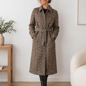 May include: A long, brown herringbone coat with a matching belt and pockets. The coat is knee-length and worn with black boots. The coat has a classic collar and a tailored fit, suitable for cooler weather.