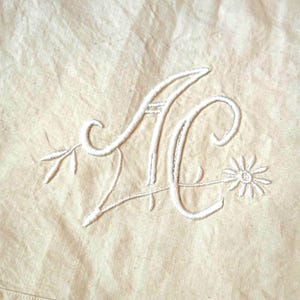 May include: Cream-colored fabric with white embroidered initials "AC" intertwined with floral and leaf accents. The embroidery is detailed, with a daisy at the end of the design. The fabric has a subtle texture.