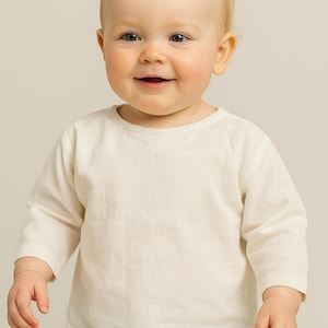 Puede incluir: Un bebé sonriente con una camisa crema de manga larga y pantalones claros. La camisa tiene un delicado ribete de encaje alrededor del cuello. El bebé tiene el pelo rubio y mira directamente a la cámara.