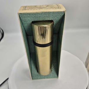 Peut inclure: Un objet cylindrique doré avec des rainures verticales, contenu dans une boîte vintage. La boîte a un intérieur vert et un dessus à motifs. Une bande noire entoure l'objet. La boîte est en carton.