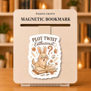 May include: A magnetic bookmark with the text "PLOT TWIST Enthusiast" and a cartoon rabbit reading a book. The bookmark is beige with a slot for the page and the Pagepal Crafts logo. The rabbit wears glasses and has a surprised expression.